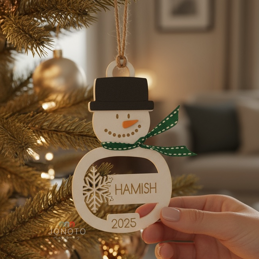 Personalized snowman ornament with 'HAMISH' and '2025' held in front of a decorated Christmas tree.